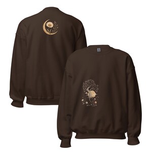 May include: A dark brown crewneck sweatshirt featuring two distinct graphic prints. The back displays a crescent moon with a daisy and foliage, while the front shows a tree with roots, a sun, and small flowers in muted beige and cream tones.