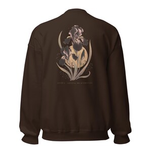 May include: A dark brown crewneck sweatshirt featuring a graphic on the back of two purple iris flowers layered over a pale yellow circle. The design includes green leaves and stems, with the text "LUPUS - LIVING BEAUTIFULLY" printed in small, light-coloured capital letters below the floral illustration.