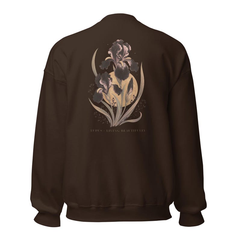 May include: A dark brown crewneck sweatshirt featuring a graphic on the back of two purple iris flowers layered over a pale yellow circle. The design includes green leaves and stems, with the text "LUPUS - LIVING BEAUTIFULLY" printed in small, light-coloured capital letters below the floral illustration.