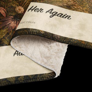 K&ouml;nnte beinhalten: Eine gefaltete Decke mit cremefarbenem Lammfellimitat-Futter und einer dunklen Blumenmuster-Au&szlig;enseite. Der Stoff zeigt den Text "Her Again" und "LECTION" in schwarzer Schrift. Das Blumenmuster enth&auml;lt braune und gr&uuml;ne botanische Elemente.