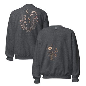 May include: A dark grey crewneck sweatshirt featuring a floral and celestial graphic. The back displays a large bouquet with a crescent moon, butterflies, and the text "WALKING BESIDE YOU." The front features a smaller, matching floral bouquet.
