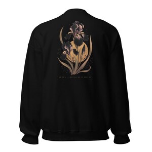 May include: A black long-sleeve sweatshirt featuring a graphic on the back of dark purple iris flowers layered over a large golden yellow circle. Below the floral design, the text reads LUPUS - LIVING BEAUTIFULLY in a small, clean font.