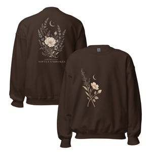 May include: A dark brown crewneck sweatshirt featuring a floral and crescent moon graphic. The back displays a large bouquet with the text "FIBROMYALGIA SOFTLY UNBROKEN," while the front features a smaller, matching floral and moon design.