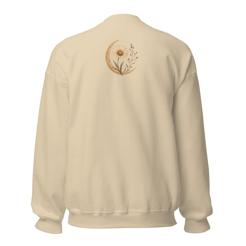 May include: A beige crewneck sweatshirt featuring a graphic on the upper back. The design depicts a crescent moon in a tan hue, centered behind a detailed illustration of a daisy flower and small sprigs of foliage in shades of brown and cream.