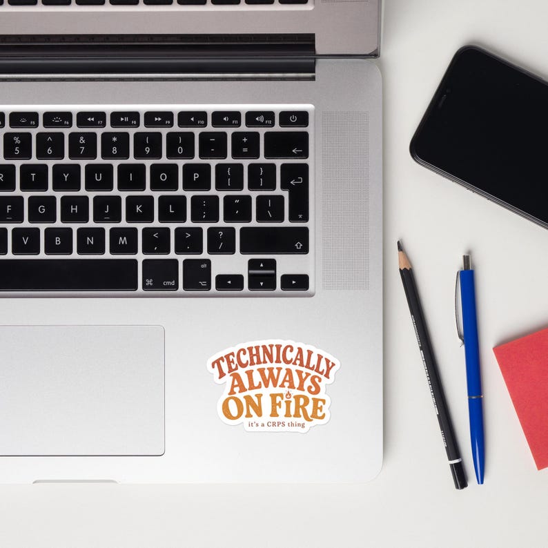 Peut inclure: Un autocollant d&eacute;coup&eacute; pr&eacute;sentant le texte orange "TECHNICALLY ALWAYS ON FIRE" avec une petite ic&ocirc;ne de flamme au-dessus du mot "ON" et le texte plus petit "it's a CRPS thing" en dessous, centr&eacute; sur le repose-poignet d'un clavier d'ordinateur portable argent&eacute;.