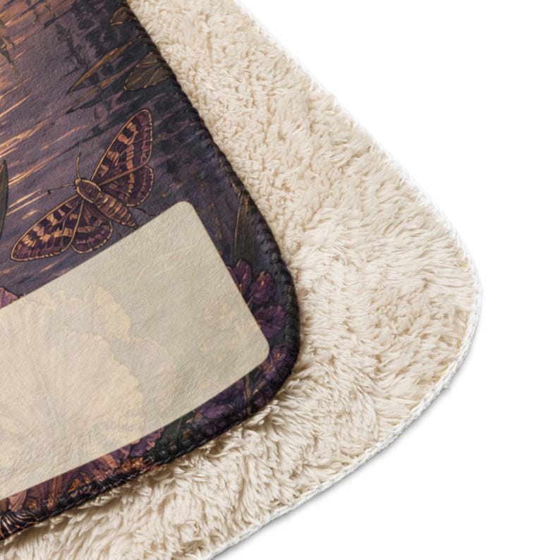 May include: A rectangular blanket featuring a dark, printed top layer with a moth and floral pattern in shades of brown, purple, and gold. The underside is made of a thick, cream-colored, textured sherpa fleece material with a soft, fuzzy appearance.