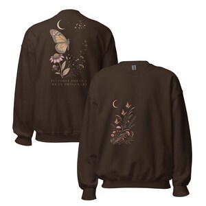 May include: A dark brown crewneck sweatshirt featuring a graphic of a butterfly, flowers, and a crescent moon. The back displays the text "INVISIBLE DOESN'T MEAN IMAGINARY" alongside a butterfly and floral design, while the front shows a smaller floral motif.