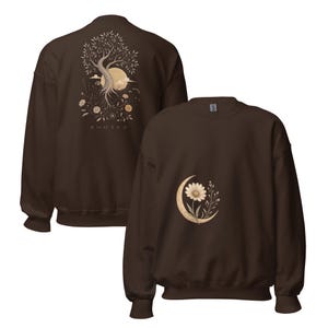 May include: A dark brown crewneck sweatshirt featuring a graphic on the back of a tree with roots, a sun, and the text "ROOTED" below it. The front of the sweatshirt displays a smaller graphic of a single daisy flower centered within a crescent moon.