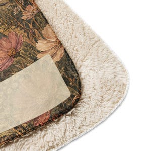 May include: A rectangular throw blanket featuring a printed floral pattern with muted pink, brown, and green botanical illustrations on one side and a thick, cream-colored, textured sherpa fleece lining on the reverse side with rounded corners.