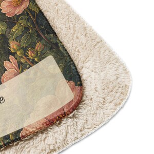 May include: A plush throw blanket featuring a dark green background with a botanical print of pink rose blossoms and green leaves. The blanket has a thick, cream-colored sherpa fleece lining on one side and a smooth, printed fabric on the other.