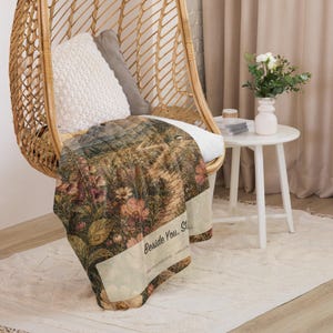 May include: A rectangular throw blanket featuring a detailed botanical print of a garden path with wildflowers, foxgloves, and greenery. The text "Beside You, Still" is printed in a dark font on a light cream-colored band across the lower section.