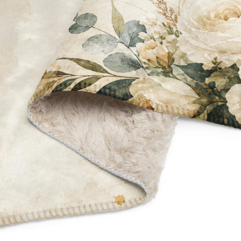May include: A soft, plush throw blanket featuring a cream-colored background with a printed floral pattern of large white roses, delicate buds, and muted green eucalyptus leaves. The underside is lined with a thick, textured beige sherpa fleece.