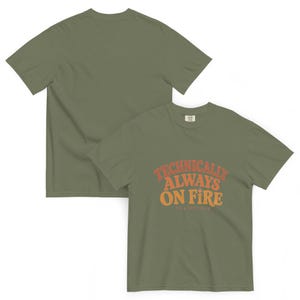 Puede incluir: Una camiseta de manga corta de color verde oliva con el texto "TECHNICALLY ALWAYS ON FIRE" en una fuente retro curva de color naranja y amarillo, con el texto m&aacute;s peque&ntilde;o "it's a good thing" impreso debajo en una fuente sans-serif clara.