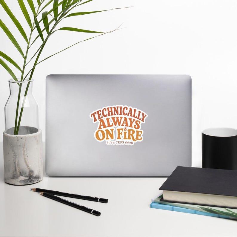 Peut inclure: Un autocollant d&eacute;coup&eacute; pr&eacute;sentant le texte "TECHNICALLY ALWAYS ON FIRE" dans une police orange audacieuse de style r&eacute;tro, avec le texte plus petit "it's a CRPS thing" en dessous. L'autocollant est appliqu&eacute; au centre d'un ordinateur portable argent&eacute; ferm&eacute;.