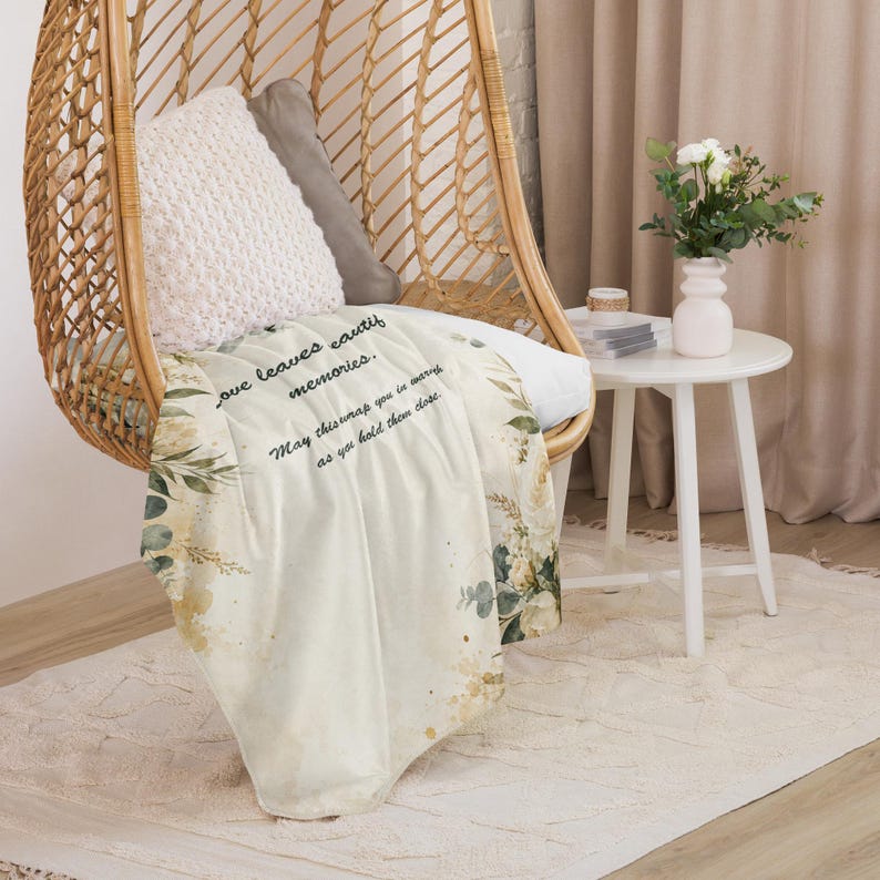 May include: A cream-colored throw blanket featuring a watercolor botanical print of green leaves and gold accents. The text reads: Love leaves beautiful memories. May this wrap you in warmth as you hold them close. The blanket is draped over a wicker chair.