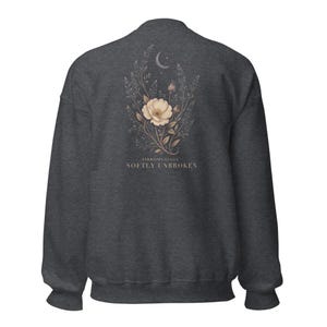 May include: A dark charcoal gray crewneck sweatshirt featuring a back graphic of a beige flower, leafy stems, and a crescent moon. Below the floral design, the text reads FIBROMYALGIA SOFTLY UNBROKEN in a clean, serif font centered on the fabric.