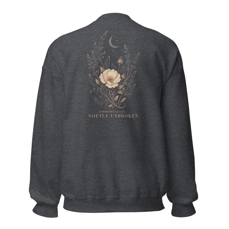 May include: A dark charcoal gray crewneck sweatshirt featuring a back graphic of a beige flower, leafy stems, and a crescent moon. Below the floral design, the text reads FIBROMYALGIA SOFTLY UNBROKEN in a clean, serif font centered on the fabric.