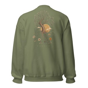 May include: An olive green crewneck sweatshirt featuring a back graphic of a tree with exposed roots, a large yellow sun, clouds, and various wildflowers. The word ROOTED is printed in small, light-colored capital letters below the illustration.