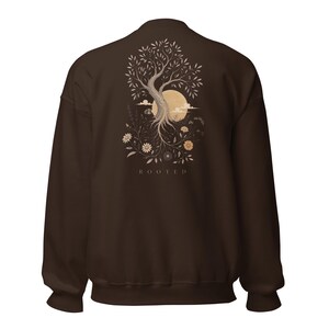 May include: A dark brown crewneck sweatshirt featuring a graphic on the back of a tree with exposed roots, surrounded by various flowers and a large circular moon. The word ROOTED is printed in capital letters below the tree illustration.
