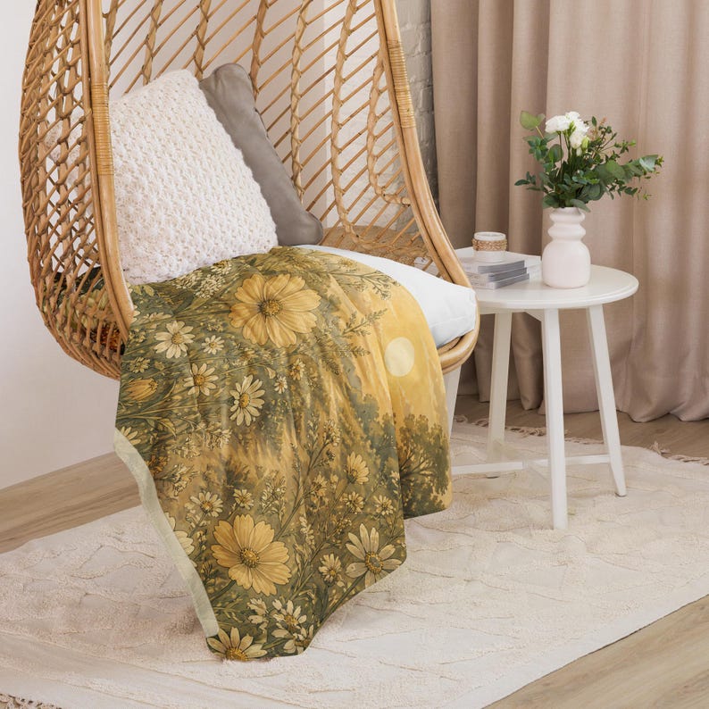 May include: A soft throw blanket featuring a detailed botanical print of yellow daisies and green foliage against a golden-hued background. The blanket is draped over a woven rattan hanging chair, showcasing a repeating floral pattern with a light border.