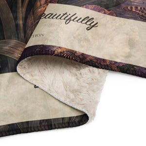 May include: A folded throw blanket featuring a dark botanical print with butterflies and flowers on one side and a cream-colored faux sherpa lining on the other. The fabric displays the text "eautifully" and "TION" in a black cursive script.
