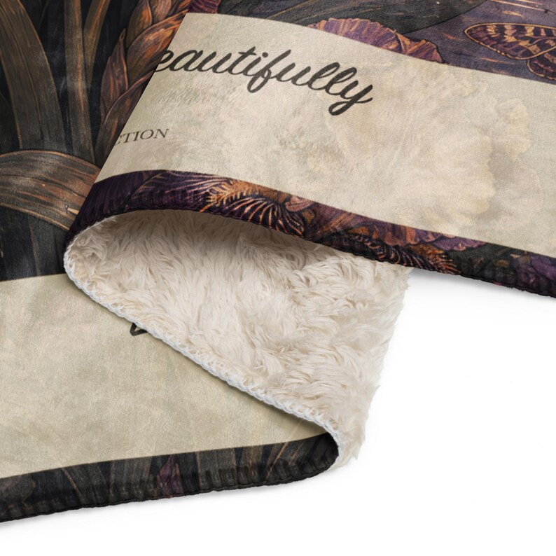 May include: A folded throw blanket featuring a dark botanical print with butterflies and flowers on one side and a cream-colored faux sherpa lining on the other. The fabric displays the text "eautifully" and "TION" in a black cursive script.