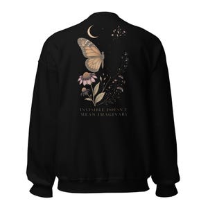 May include: A black long-sleeve sweatshirt featuring a graphic on the back. The design includes a monarch butterfly, a crescent moon, and floral elements in muted tones. Below the illustration, the text reads: INVISIBLE DOESN'T MEAN IMAGINARY.