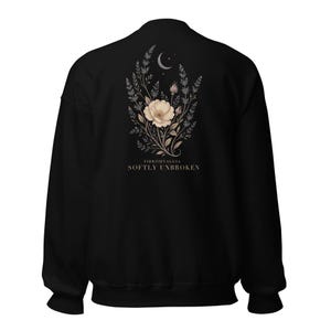 May include: A black long-sleeve sweatshirt featuring a central graphic on the back. The design displays a beige flower surrounded by botanical sprigs and a crescent moon. Below the illustration, the text reads FIBROMYALGIA SOFTLY UNBROKEN.