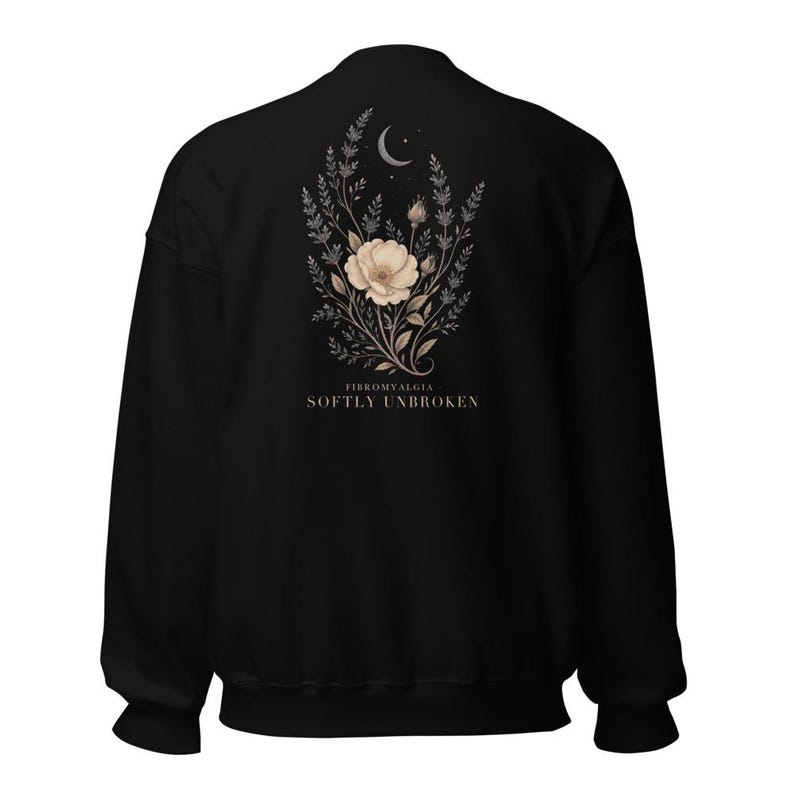 May include: A black long-sleeve sweatshirt featuring a central graphic on the back. The design displays a beige flower surrounded by botanical sprigs and a crescent moon. Below the illustration, the text reads FIBROMYALGIA SOFTLY UNBROKEN.