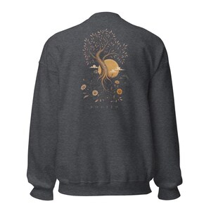 May include: A dark charcoal gray crewneck sweatshirt featuring a graphic on the back. The design depicts a tree with exposed roots, a large yellow sun, clouds, and various flowers. The word ROOTED is printed in small capital letters below the graphic.