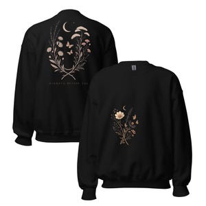 May include: A black crewneck sweatshirt featuring a floral and celestial graphic. The back displays a large bouquet with a crescent moon, butterflies, and the text "WALKING BESIDE YOU." The front shows a smaller, matching floral and moon design.