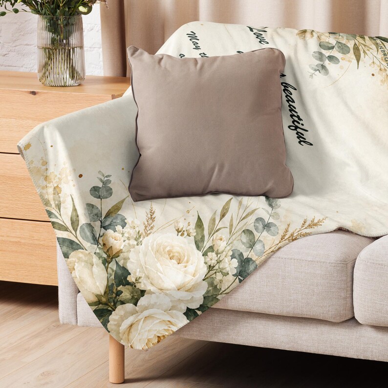 May include: A cream-colored throw blanket featuring a watercolor floral print of white roses and eucalyptus leaves, with black cursive text that reads Love, May the, and beautiful, draped over a sofa with a solid taupe square throw pillow.