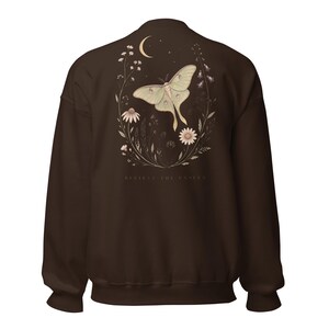 Może przedstawiać: Ciemnobrązowa bluza z długim rękawem z nadrukiem na plecach przedstawiającym jasnozieloną ćmę otoczoną polnymi kwiatami i p&oacute;łksiężycem. Poniżej ilustracji znajduje się napis "BELIEVE THE UNSEEN" wykonany małymi, jasnymi, wielkimi literami.