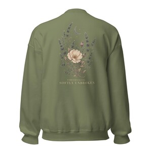 May include: An olive green crewneck sweatshirt featuring a back graphic of a floral arrangement with lavender sprigs, a central pale flower, and a crescent moon. Below the illustration, the text reads FIBROMYALGIA SOFTLY UNBROKEN.