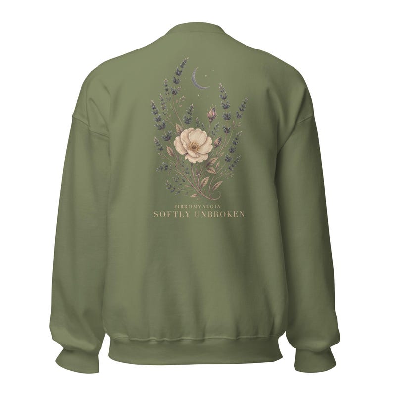 May include: An olive green crewneck sweatshirt featuring a back graphic of a floral arrangement with lavender sprigs, a central pale flower, and a crescent moon. Below the illustration, the text reads FIBROMYALGIA SOFTLY UNBROKEN.