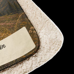 K&ouml;nnte beinhalten: Eine Decke mit Sherpa-Futter und einem aufgedruckten Landschaftsmotiv mit Gebirge, Tannen und einer Blumenwiese. Unten links befindet sich ein cremefarbener, rechteckiger Aufn&auml;her mit dem teilweise sichtbaren schwarzen Schriftzug in.