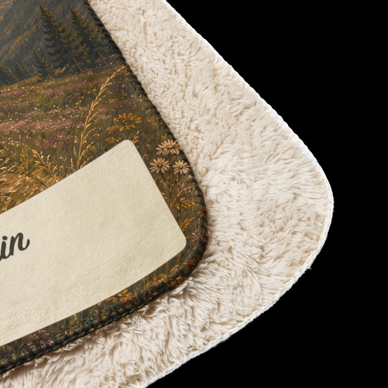 K&ouml;nnte beinhalten: Eine Decke mit Sherpa-Futter und einem aufgedruckten Landschaftsmotiv mit Gebirge, Tannen und einer Blumenwiese. Unten links befindet sich ein cremefarbener, rechteckiger Aufn&auml;her mit dem teilweise sichtbaren schwarzen Schriftzug in.