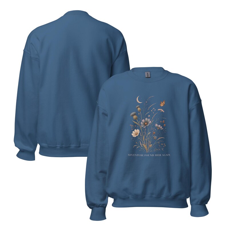 May include: A blue crewneck sweatshirt featuring a graphic print of wildflowers, a crescent moon, and a butterfly. The text ADVENTURE FOUND HER AGAIN is printed below the floral design. The image displays both the front and back of the garment.