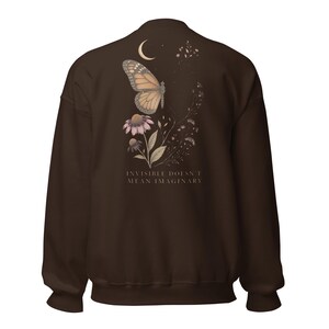May include: A dark brown long-sleeve sweatshirt featuring a back graphic of a monarch butterfly, a crescent moon, and wildflowers. Below the illustration, the text reads: INVISIBLE DOESN'T MEAN IMAGINARY. The design uses muted earth tones.
