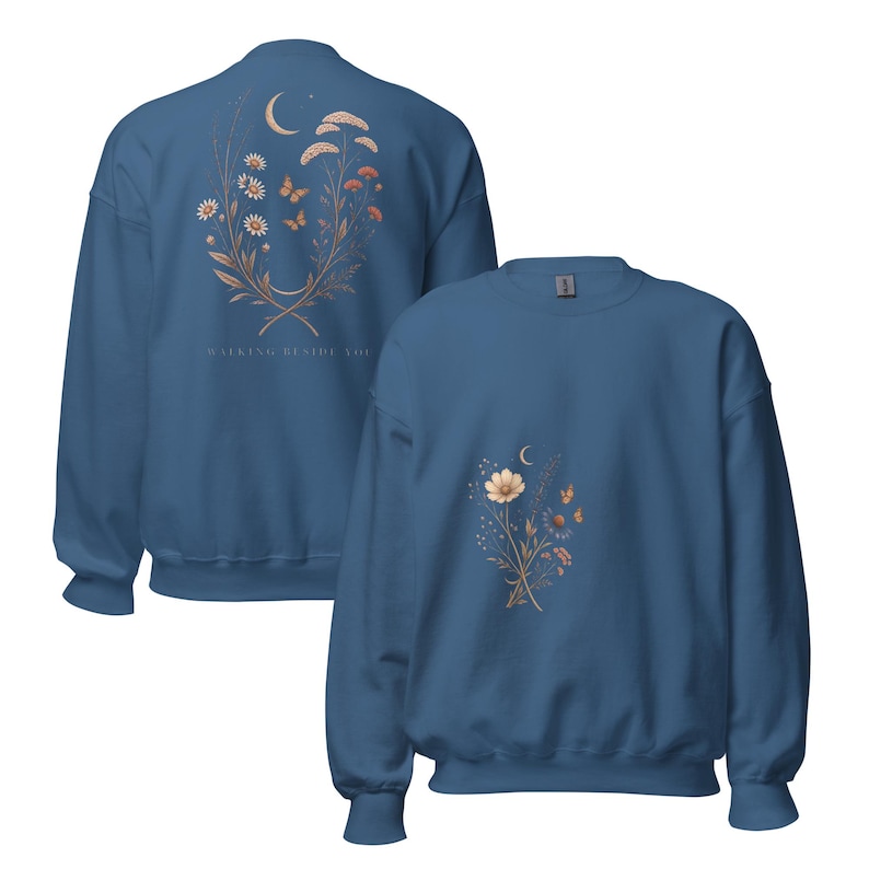 May include: A blue crewneck sweatshirt featuring a floral and crescent moon graphic on the front and a larger matching design on the back with the text "WALKING BESIDE YOU". The illustrations include daisies, wildflowers, and butterflies in muted tones.