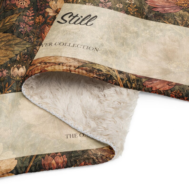 May include: A plush throw blanket featuring a dark botanical print with muted orange, brown, and green floral patterns. The text Still FLOWER COLLECTION and THE O is visible on the fabric. The underside is a thick, cream-colored sherpa fleece.
