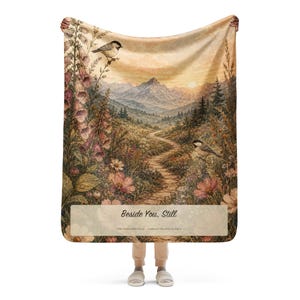 May include: A rectangular throw blanket featuring a landscape illustration of a winding stone path leading toward mountains at sunset. The scene includes foxglove flowers, small birds, and the text "Beside You, Still" and "THE ONES WHO STAY CAREGIVER COLLECTION".