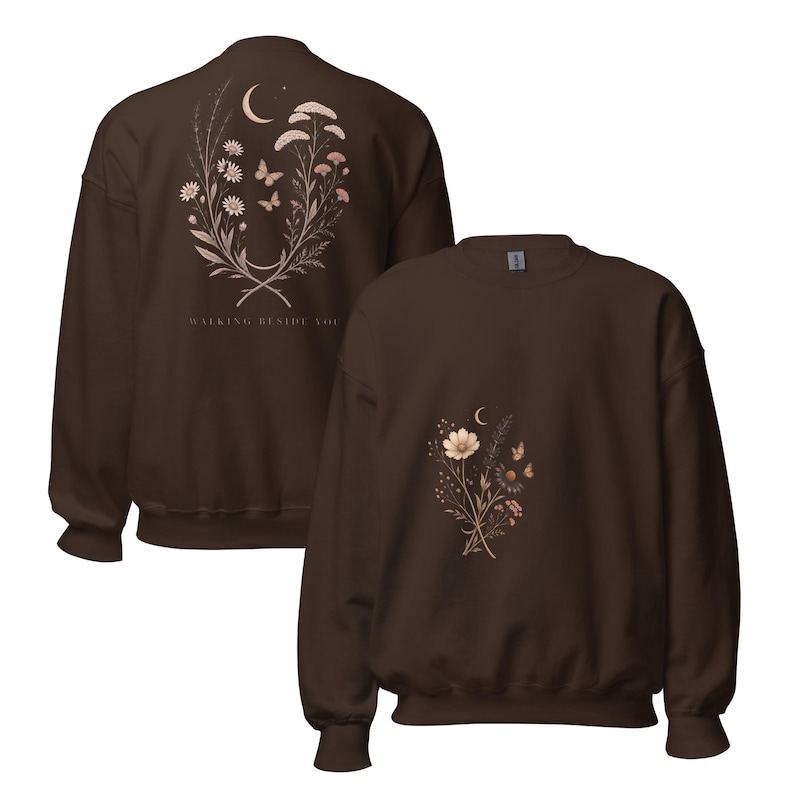 May include: A dark brown crewneck sweatshirt featuring a floral and celestial graphic. The back displays a bouquet of wildflowers, butterflies, and a crescent moon with the text WALKING BESIDE YOU. The front features a smaller matching floral design.