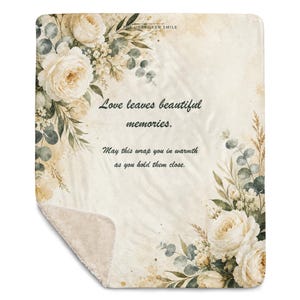 May include: A rectangular throw blanket with a cream-colored background featuring watercolor-style illustrations of white roses and eucalyptus leaves. The text reads: Love leaves beautiful memories. May this wrap you in warmth as you hold them close.
