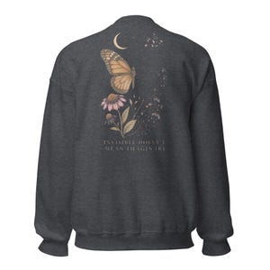 May include: A dark charcoal gray crewneck sweatshirt featuring a back graphic of a monarch butterfly, a crescent moon, and purple coneflower blossoms. The text "INVISIBLE DOESN'T MEAN IMAGINARY" is printed in a light, serif font below the floral design.