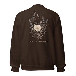 May include: A dark brown sweatshirt featuring a back graphic of a white flower, lavender sprigs, and a crescent moon. Below the floral design, the text reads FIBROMYALGIA SOFTLY UNBROKEN in a clean, light-colored serif font centered on the fabric.