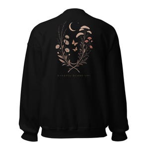 May include: A black long-sleeve sweatshirt featuring a back graphic of a crescent moon, two butterflies, and various wildflowers in muted earth tones. Below the floral arrangement, the text "WALKING BESIDE YOU" is printed in a simple font.