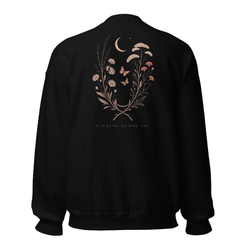 May include: A black long-sleeve sweatshirt featuring a back graphic of a crescent moon, two butterflies, and various wildflowers in muted earth tones. Below the floral arrangement, the text "WALKING BESIDE YOU" is printed in a simple font.