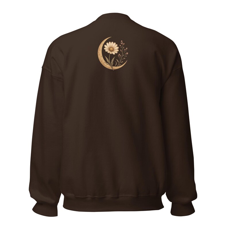 May include: A dark brown long-sleeve sweatshirt featuring a graphic on the upper back. The design depicts a beige crescent moon cradling a single daisy flower and delicate sprigs of small wildflowers, rendered in a soft, muted color palette.