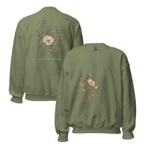 May include: An olive green crewneck sweatshirt featuring a floral graphic with a crescent moon. The back displays a larger bouquet with the text "FIBROMYALGIA SOFTLY UNBROKEN" below it, while the front shows a smaller, matching floral design.